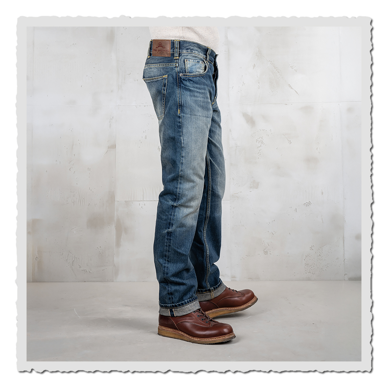 Seitliche Ansicht einer getragenen blauen Jeans mit hellen Waschungen und braunen Schuhen vor heller Studiowand.